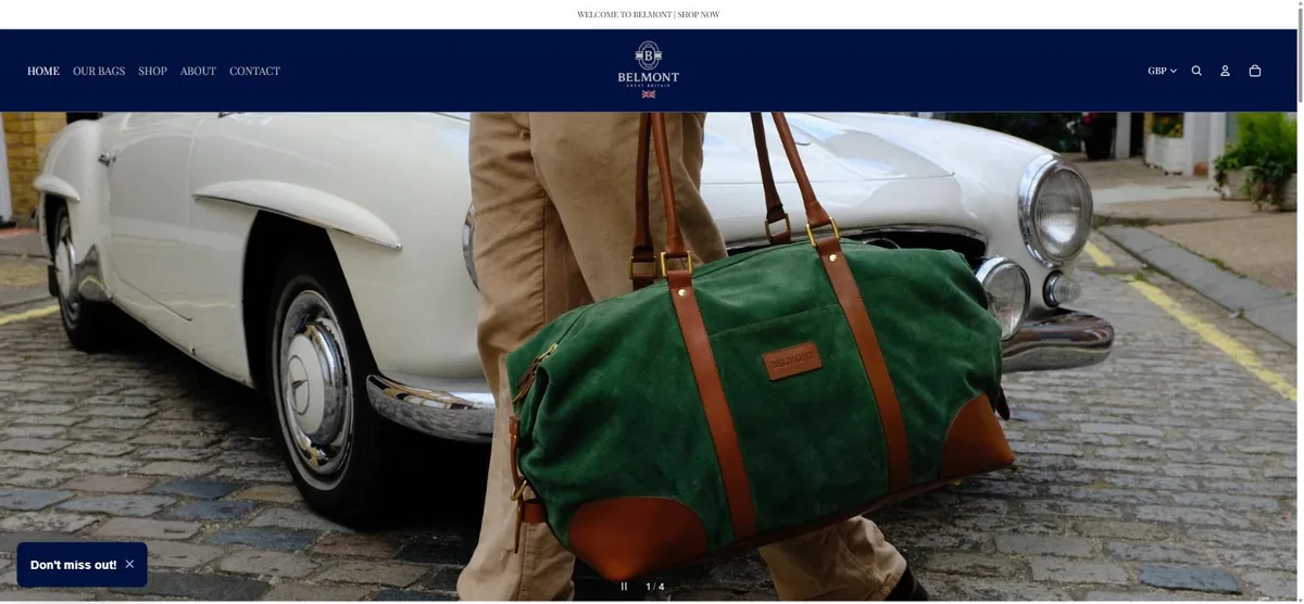 Belmont GB homepage: navy header above a lifestyle photograph of a green suede holdall being carried past a classic car.